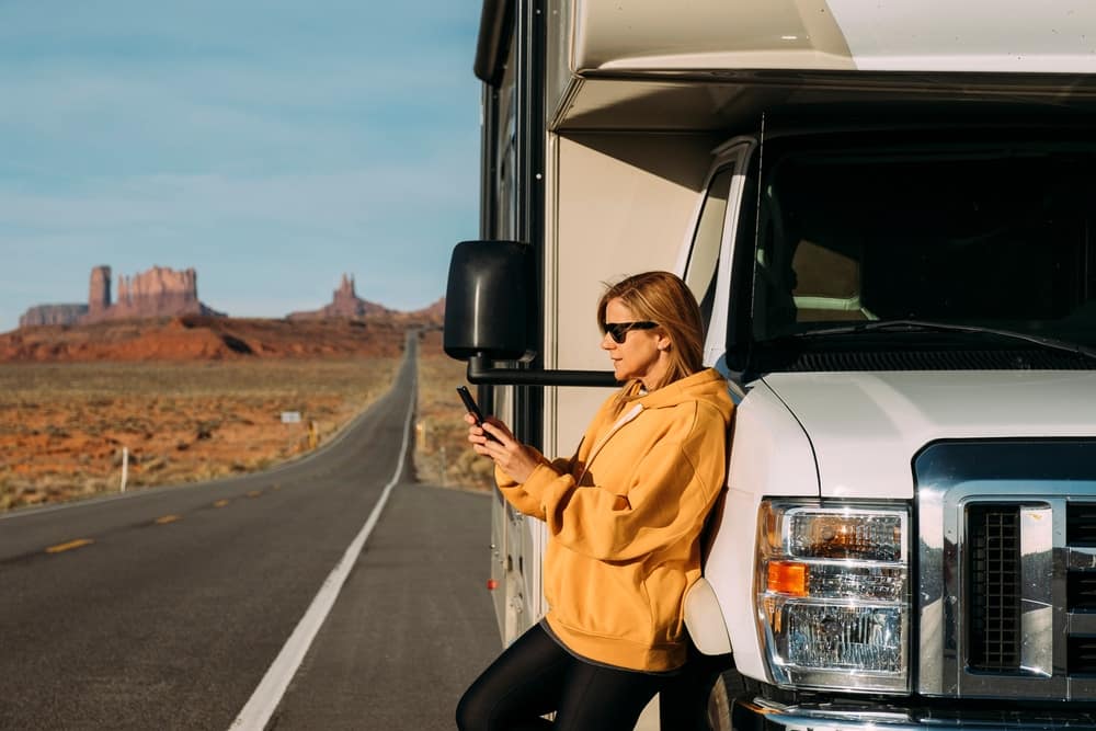 A woman standing next to an RV, browsing her phone during camping season.