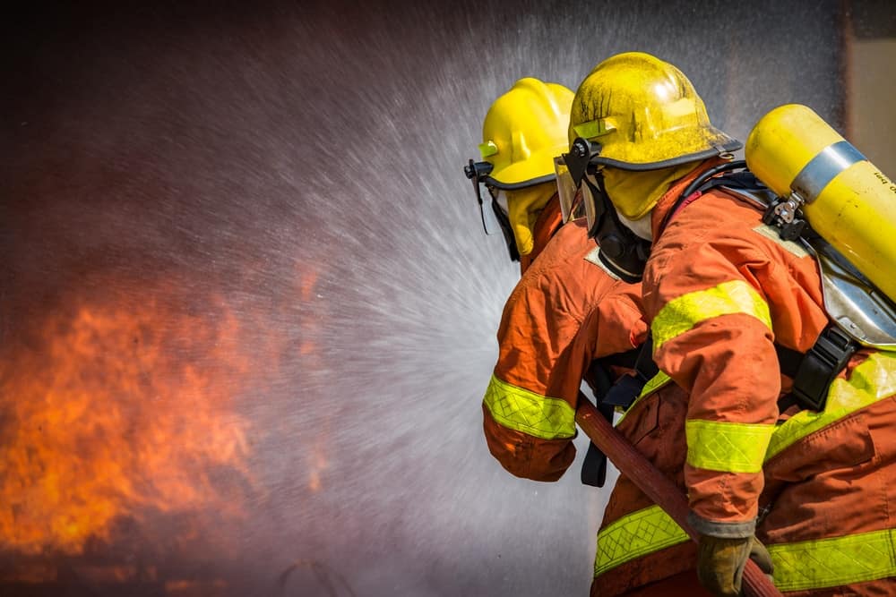 Two firefighters evacuating residents from a wildfire incident in Alberta and spraying water in front of an unprecedented levels of fire.