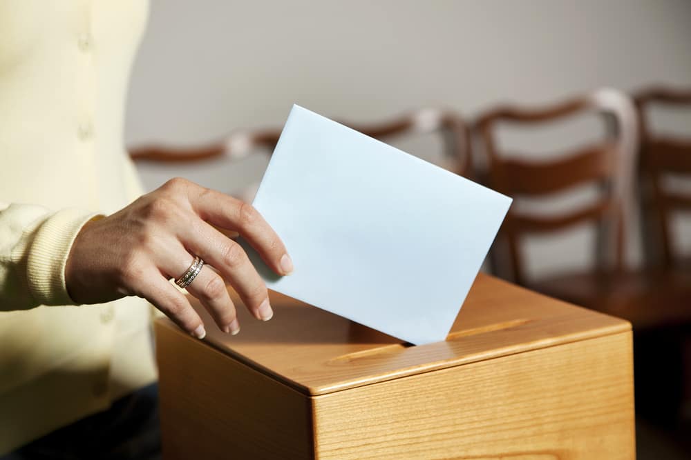 In Otsego Lake Park, a person is putting a blue envelope into a wooden voting box during the tie vote for campsite improvements.