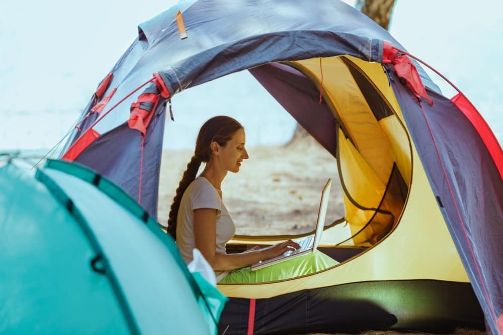 A woman enjoying camp life to the fullest, ensuring 100% uptime as she sits comfortably in a tent with her laptop.