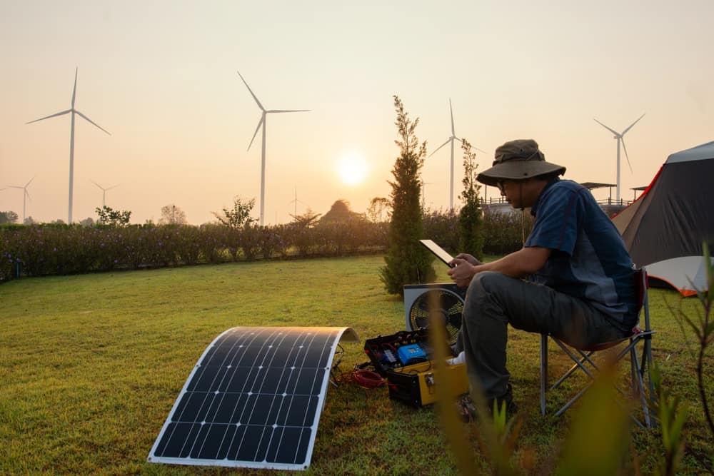 A man is working on a solar panel in front of wind turbines as a part of eco-friendly practices.