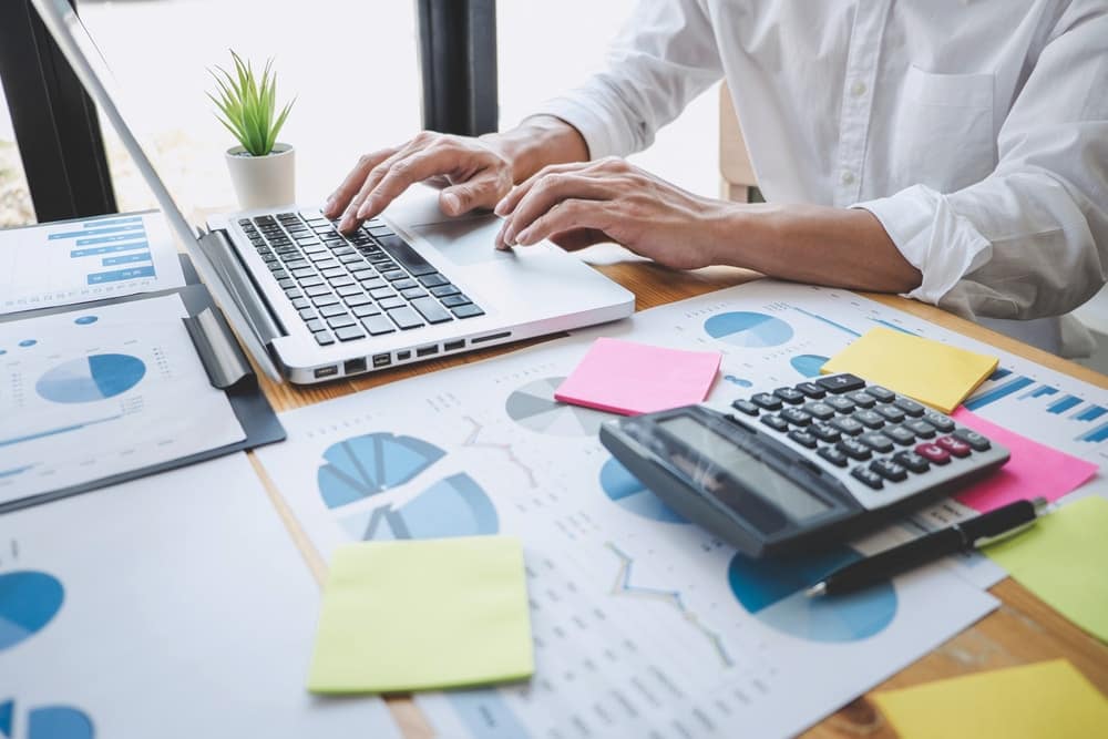A man utilizing the IDeaS Revenue Management System on a laptop at a desk, surrounded by papers and a calculator.