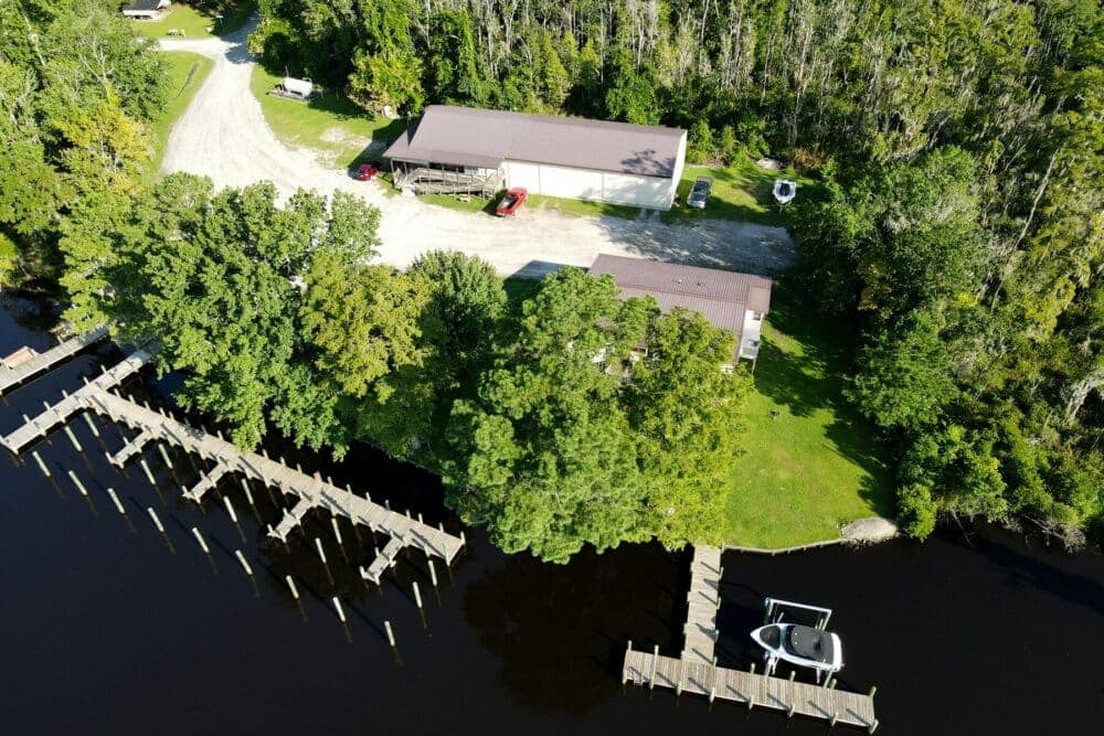 Aerial view of a rural property with a house, large shed, dock, and multiple boats at Cotton Patch Landing, surrounded by trees near a water body.