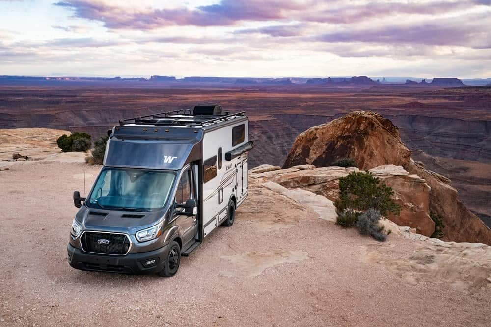 A motorhome, seemingly on auto draft, is parked on a cliff overlooking a vast canyon and mountainous landscape under a partly cloudy sky.