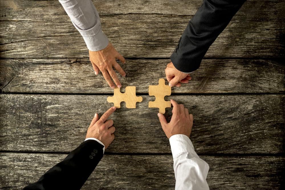 Three business people holding puzzle pieces together on a wooden table as they acquire a Travel Tech app for the Camping Community.