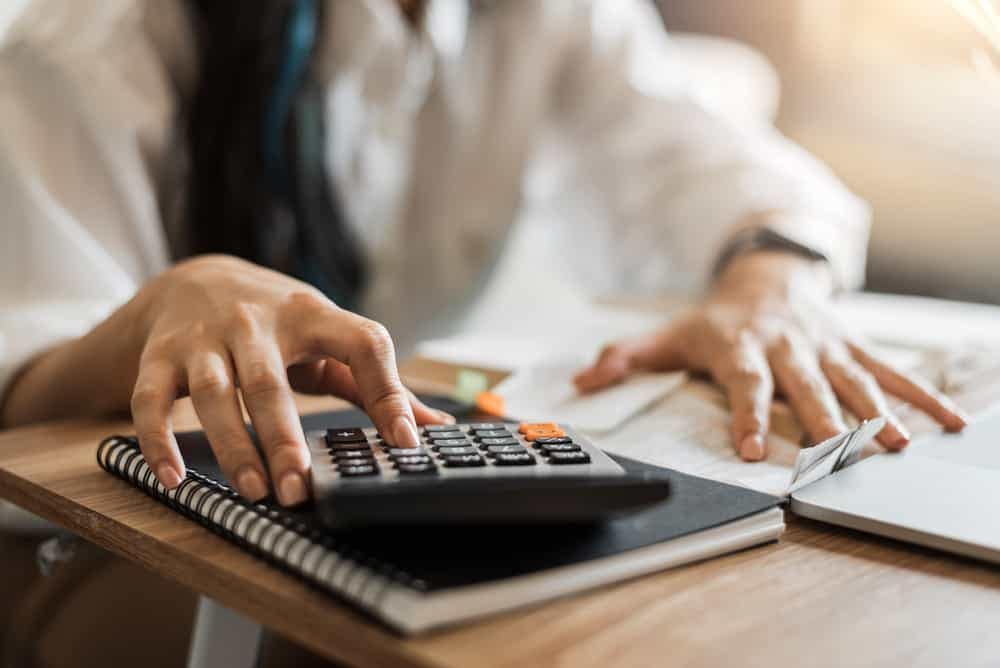 A woman is using a calculator on a desk while pondering the potential consequences of interest hikes and inflation, foreseeing tough months ahead.