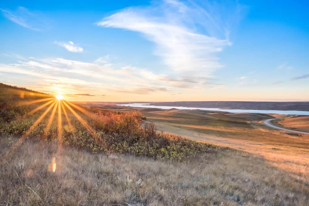 The sun rises over a grassy field in Saskatchewan, with a lake in the background.
