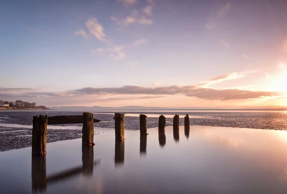 The sun is setting over a beach with wooden posts in the water at a Glamping Site.