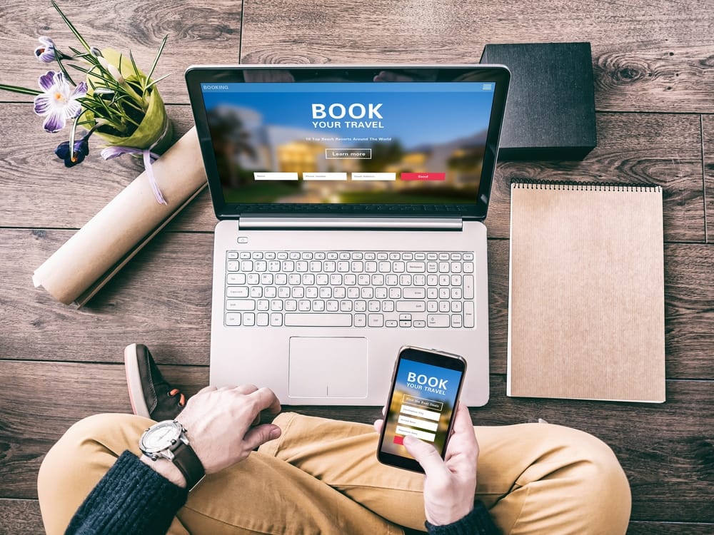 A man is using a laptop and a cell phone to book a hotel.