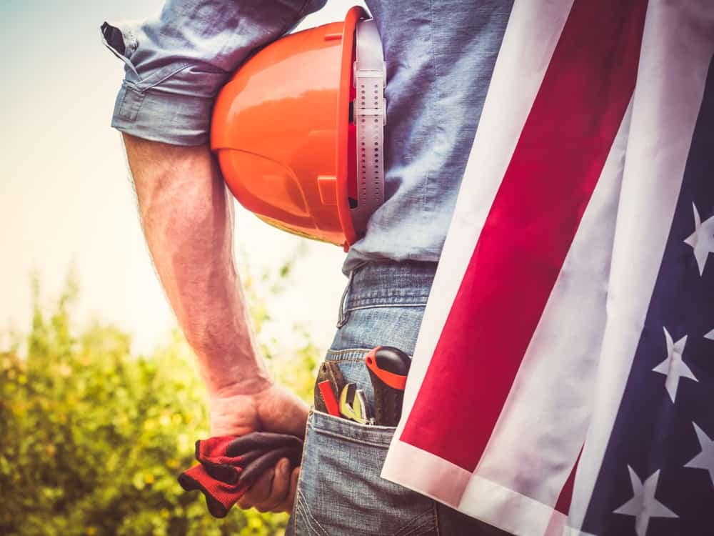 A man with a hard hat and tools proudly holding an American flag at a Labor Day celebration, surrounded by cabins in picturesque Texas campsites.