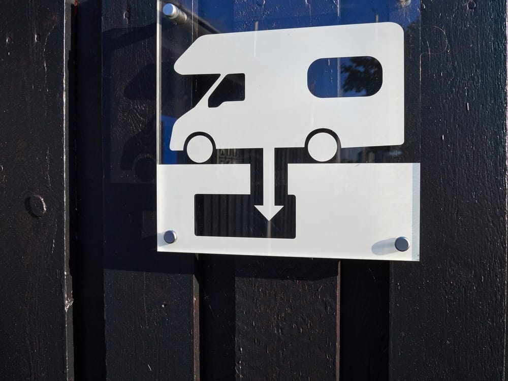 A sign with an RV on it attached to a black fence indicating the location of a Sani-Dump Station in Vernon.