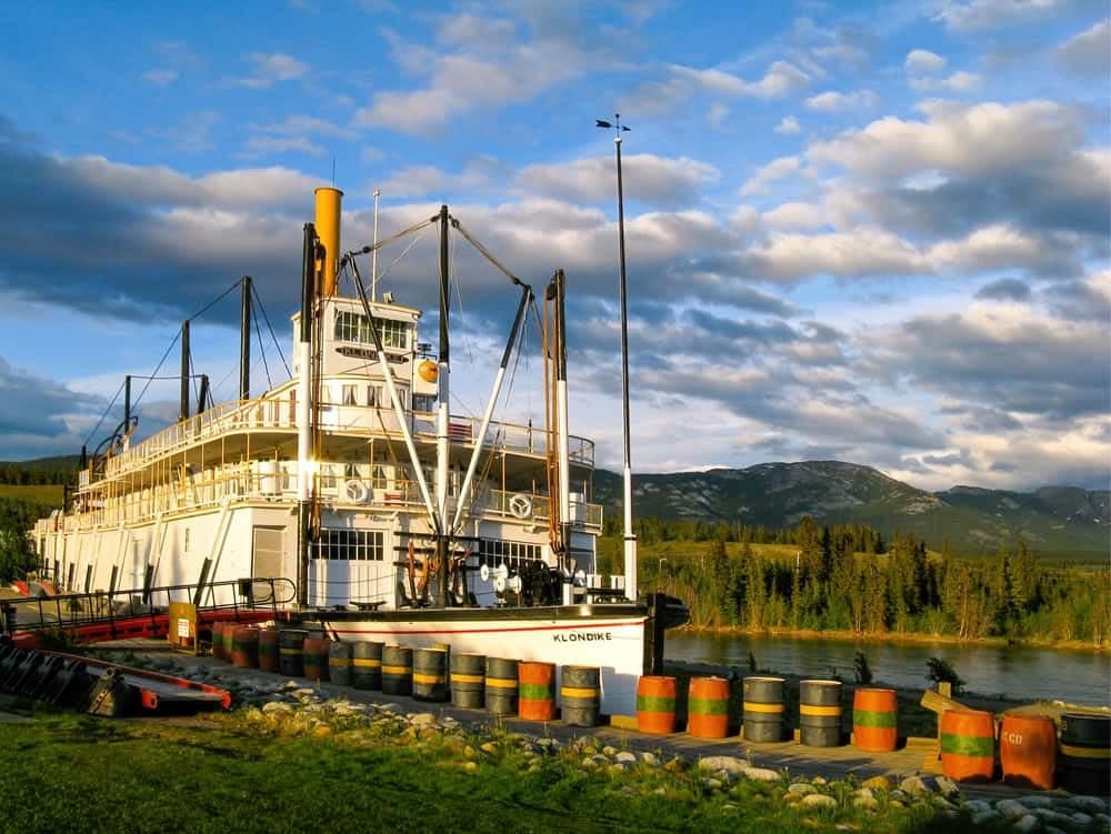 A S.S. Klondike steamboat docked at a river with majestic mountains in the background, showcasing its significance as a National Historic Site.