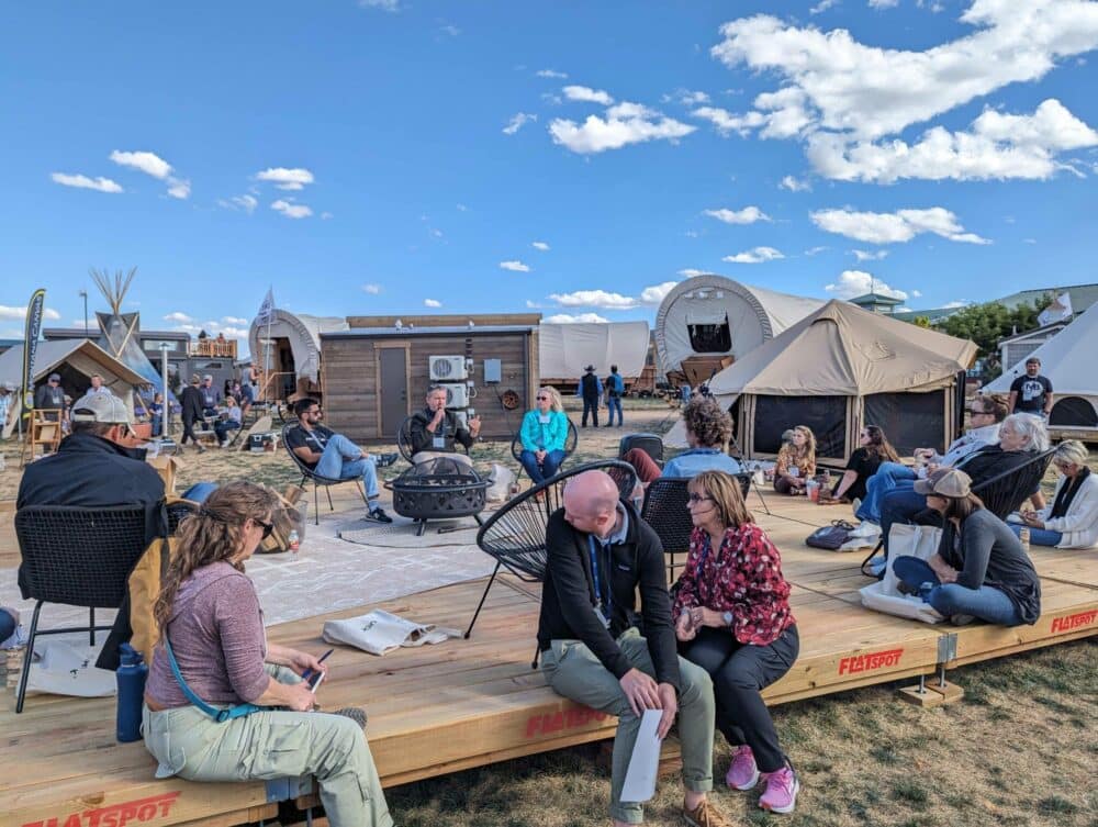 A group of people sitting on a wooden deck in front of tents at the Glamping Show Americas 2023, engaging with Exhibitor Presentations and discussing the latest developments in the Glamping