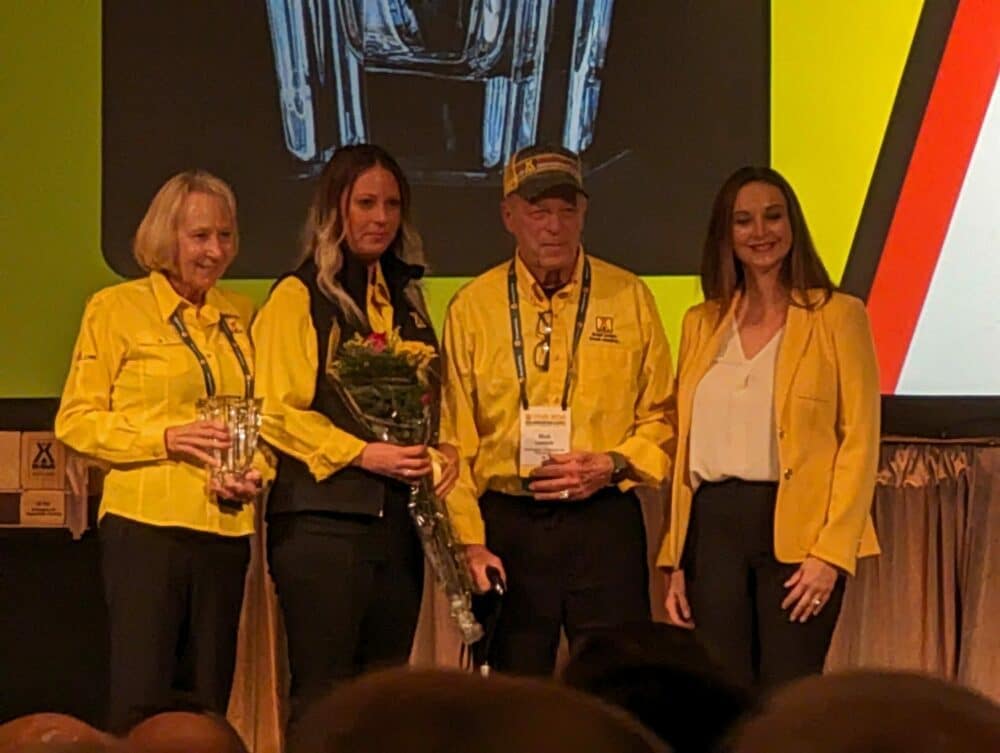 A group of Long-Time Partners standing in front of a stage adorned with flowers, proudly receiving the Founder's and President's Awards from KOA.