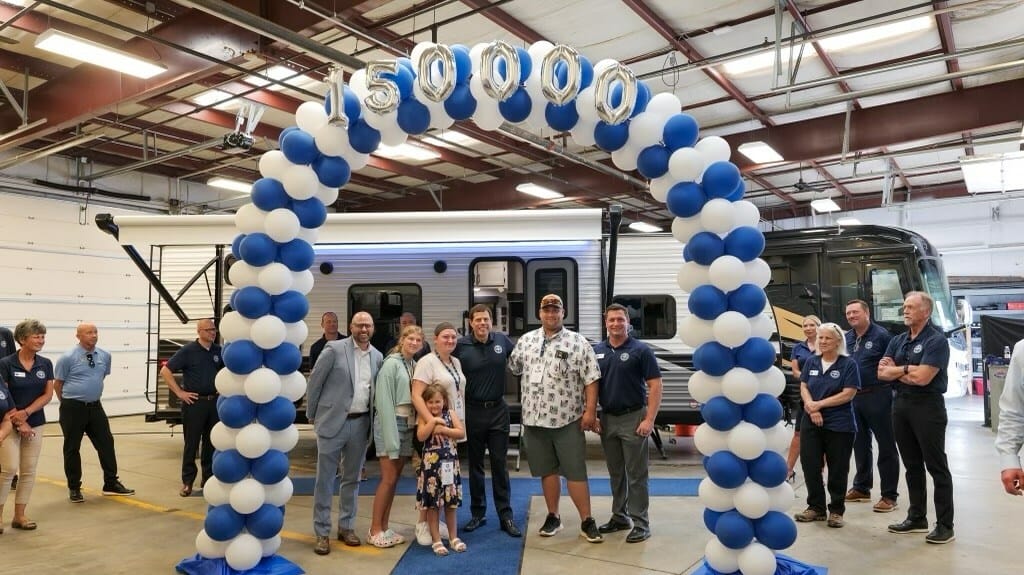 A group of people standing in front of a Blue Compass RV, celebrating the industry record of 150,000 RV sales.