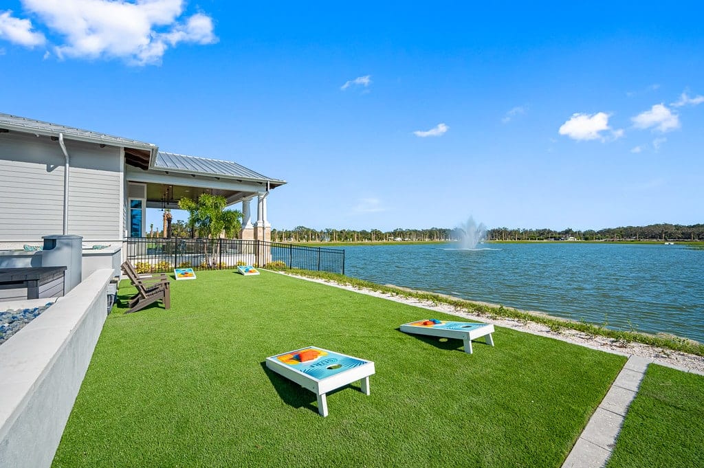 A lakeside yard featuring a game area with multiple cornhole boards, a modern house with large windows to the left, and a lake with a fountain in the background, reminiscent of the finest RV parks.