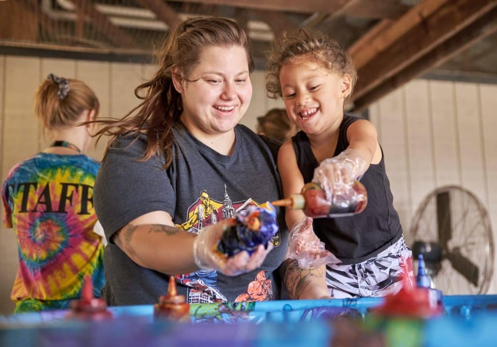 A woman and a young girl smiling while doing Mother's Day activities, using colorful paints at a workshop table, with another person in the background.