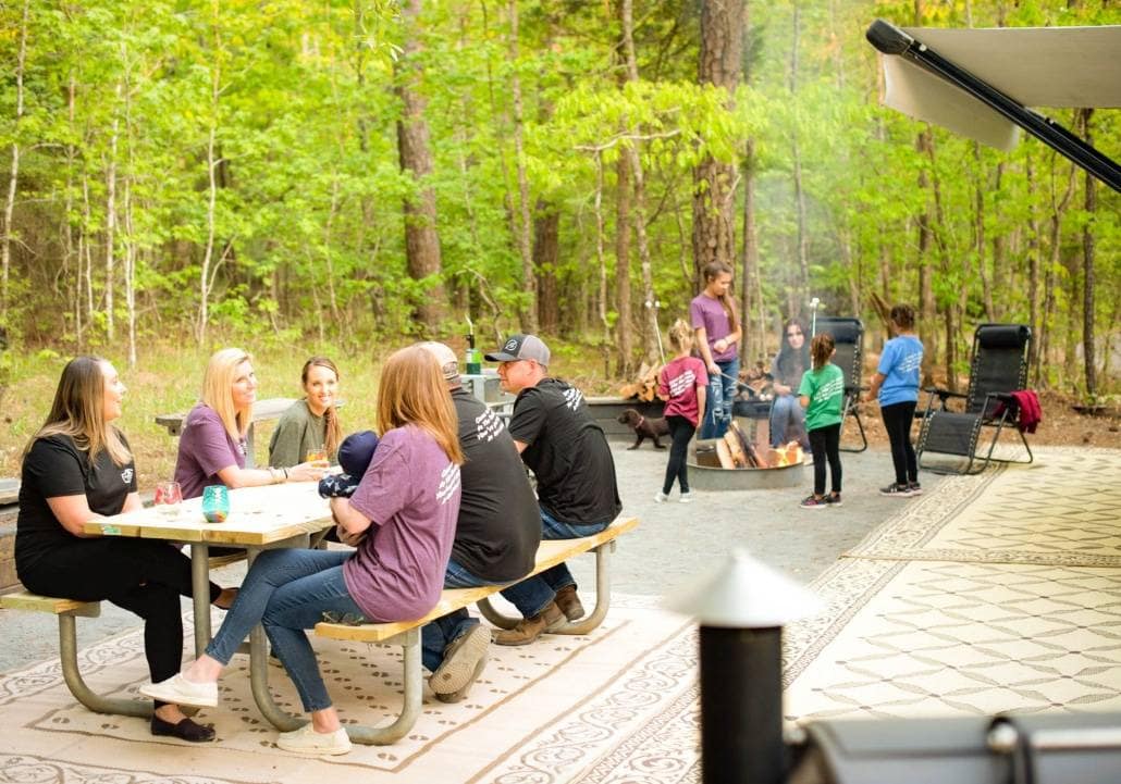 A community of people, including women, gathered around a picnic table in the woods of South Carolina.