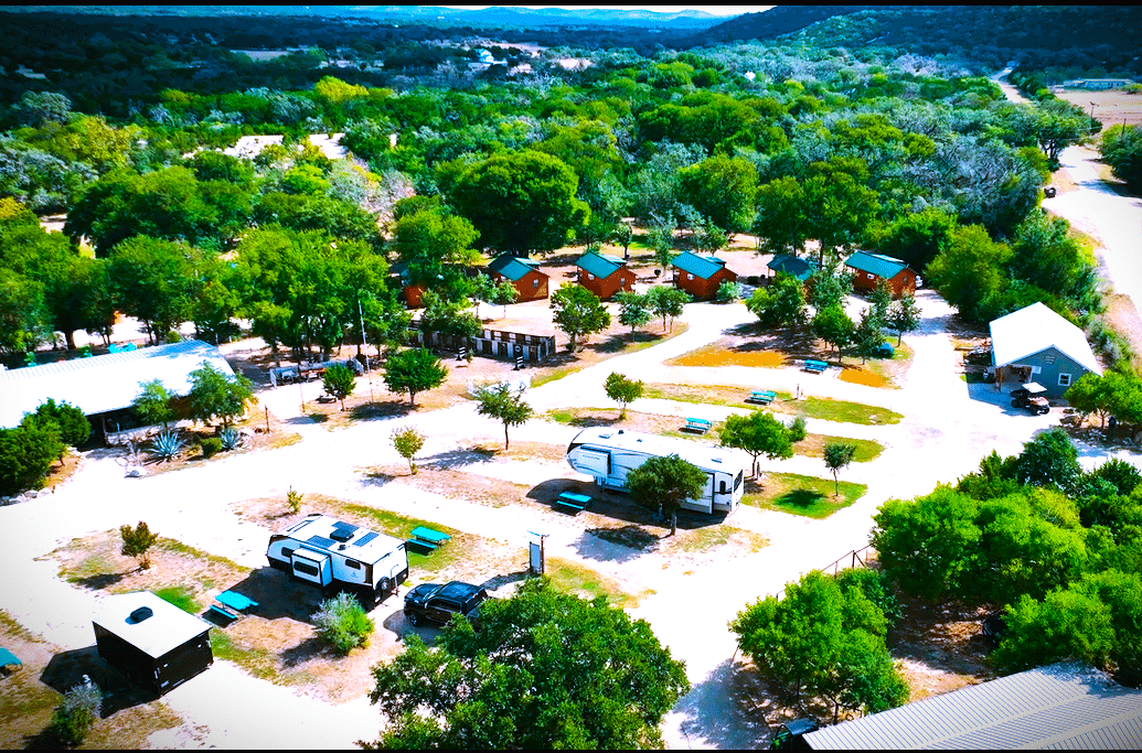 An auto draft of an aerial view captures a campground with several RVs, cabins, and tents nestled among trees and dirt roads, all surrounded by rolling green hills in the background.