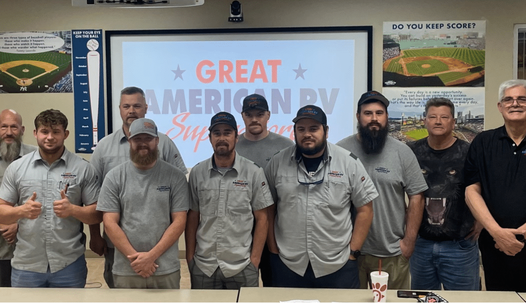 Group of men standing in front of a screen displaying "great american rv superstore" in a room decorated with baseball-themed posters and an Auto Draft poster.