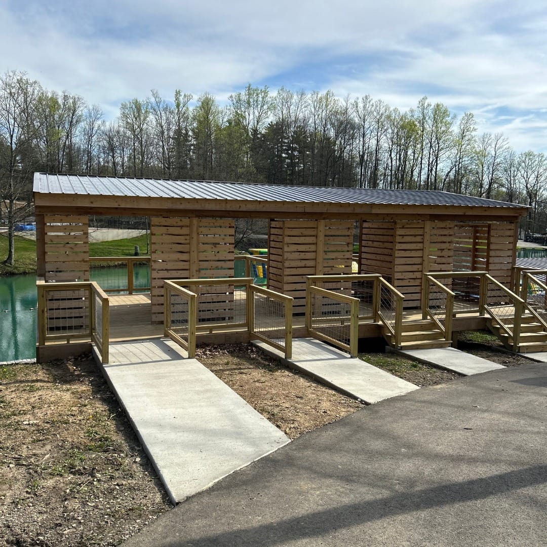 A wooden shelter with a metal roof and multiple ramps is positioned by a pond, surrounded by trees, offering a touch of luxury glamping near Yogi Bear’s Jellystone Park.