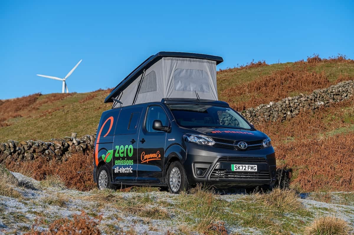 A Low-Emission campervan parked on a hill with wind turbines in the background.