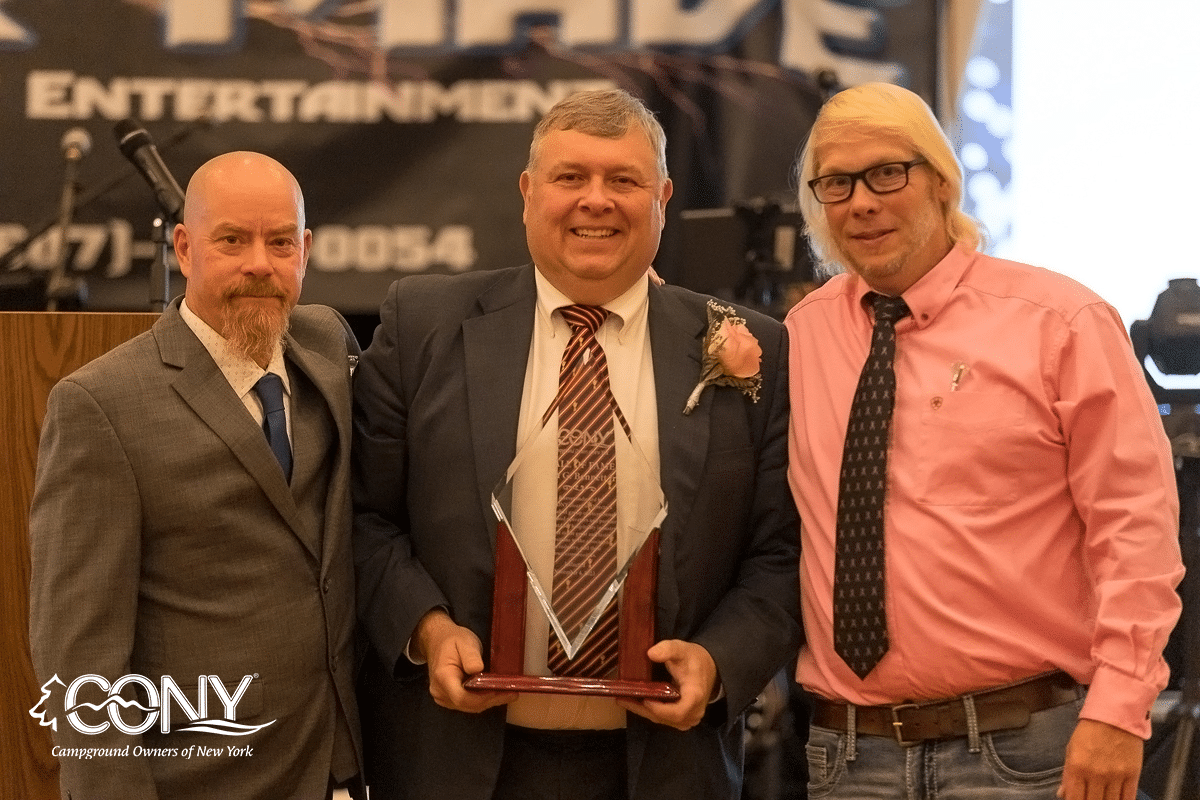 Three men, including Bennett, posing with an award in front of a stage at the Campground Owners of New York Hall of Fame.
