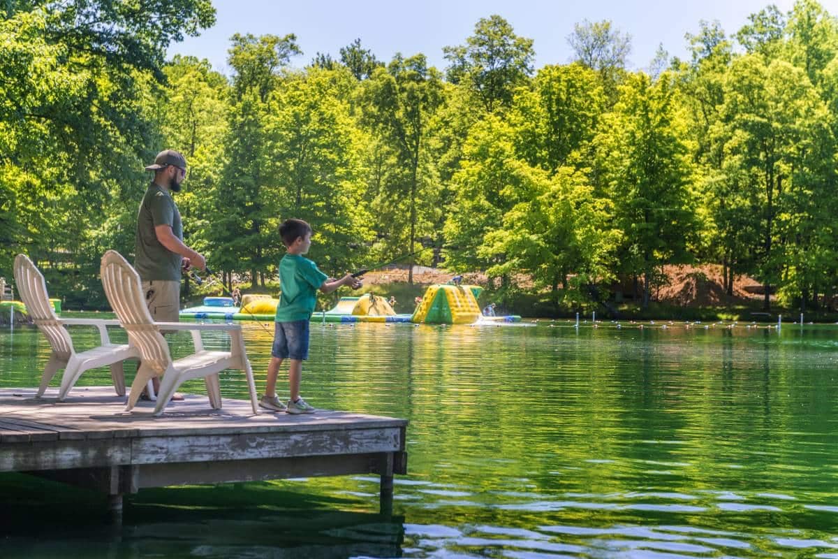 An adult and child fishing from a dock on an Auto Draft sunny day with recreational water equipment in the background.