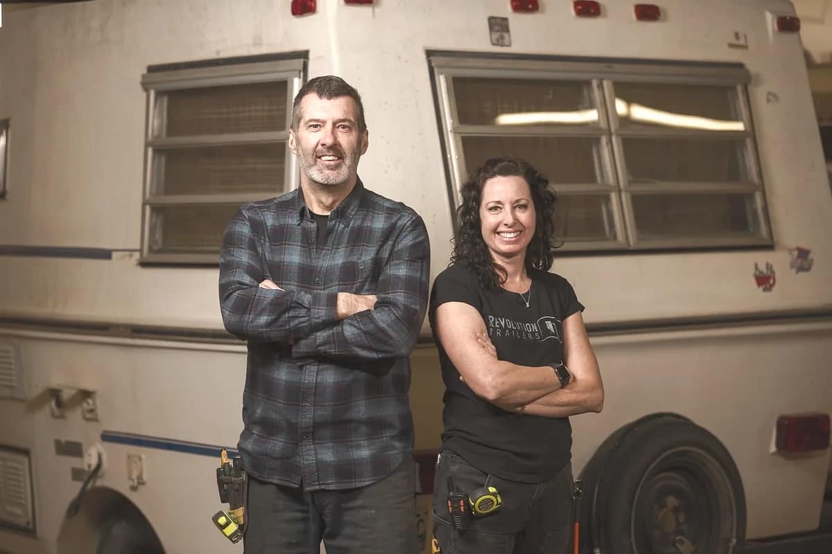 A man and woman smiling, standing confidently with arms crossed in an auto workshop, in front of a camper van. Both are dressed in casual work attire.