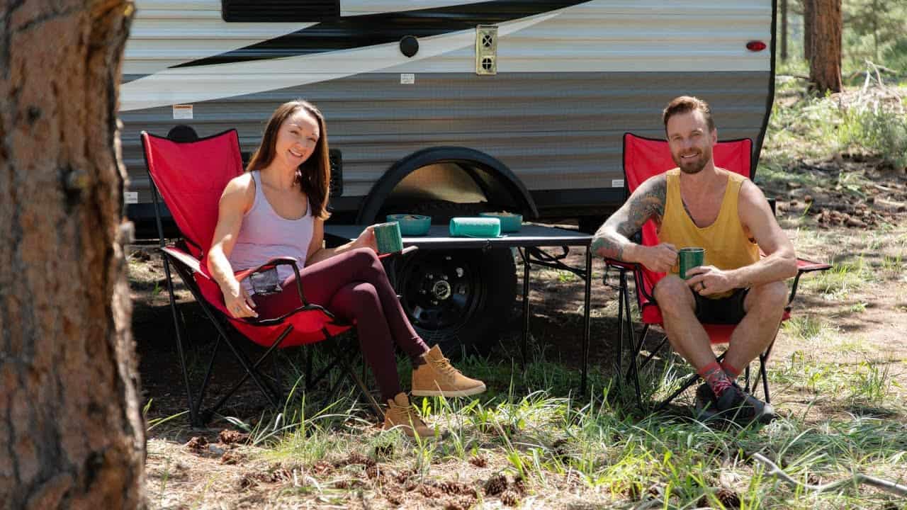 A man and woman sitting in front of an rv in the woods.