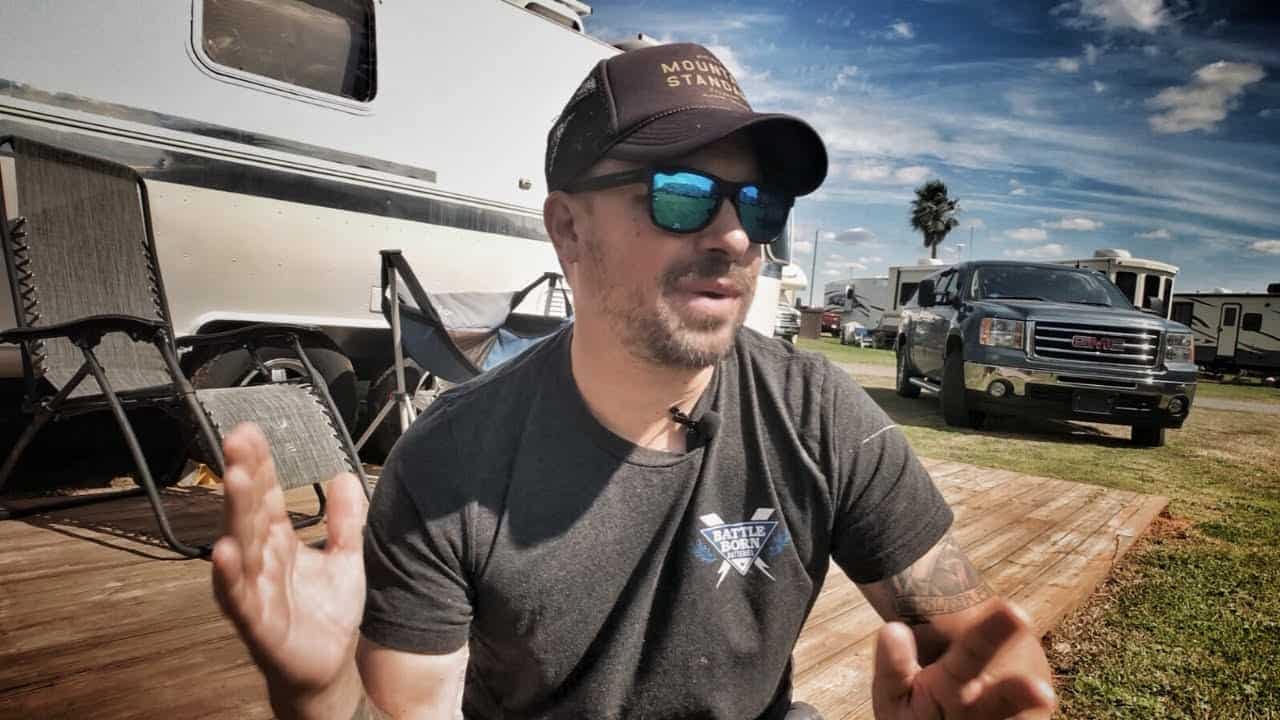 A man wearing sunglasses and a hat is sitting on a deck in front of an RV, avoiding bad RV parks.