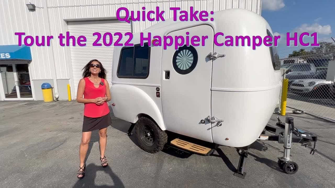 A woman standing in front of a trailer with the words RV Tour the 2021 happier camper hc.