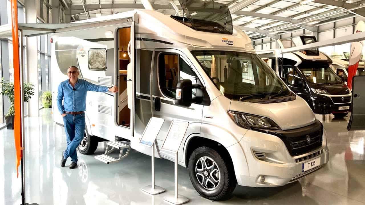 A man standing next to a white RV in a showroom, showcasing the Carado T135.