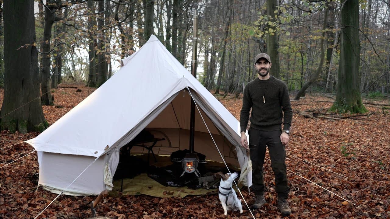 A man and his dog standing next to a canvas tent in the woods during early winter camping.
