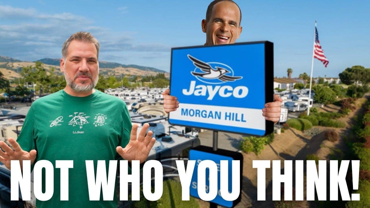 Two men holding up a sign that says "Jayco - not who you think" at an RV dealership.