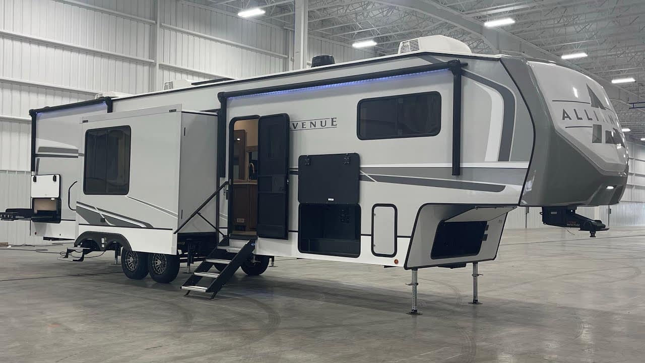 A white and gray 2024 Alliance Avenue 38DBL rv with a toy hauler design is parked in a large warehouse.