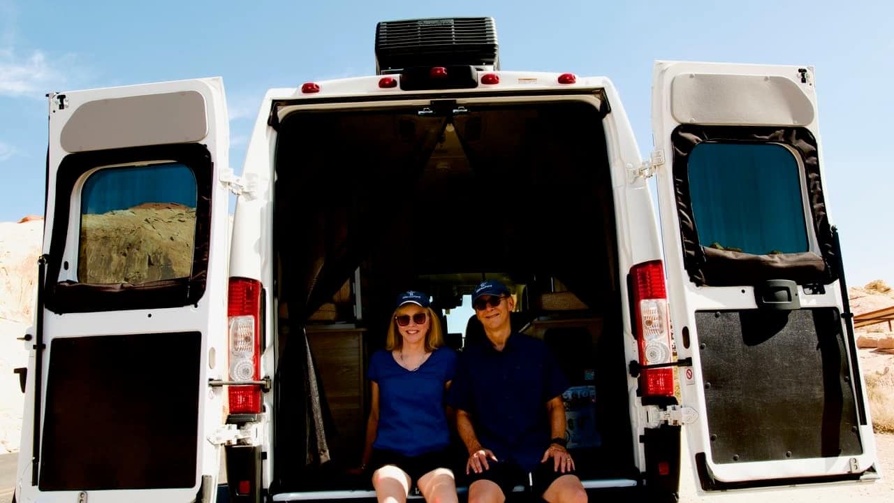 A man and a woman sit in the back of an open van with the doors ajar. They are both wearing sunglasses and blue shirts. The van, part of the Blacksford RV Rentals fleet, appears to be parked in a desert area with rocky terrain.
