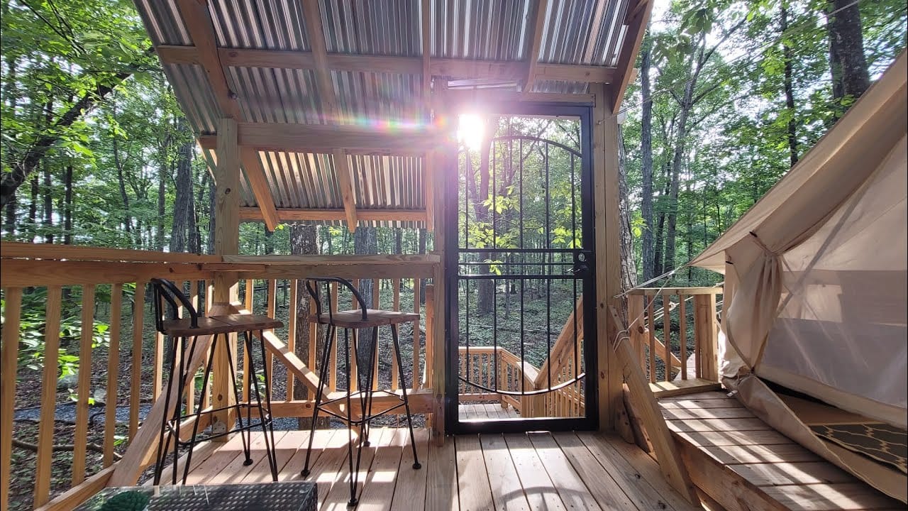 A wooden deck in a forest with bar stools, a table, and a tent. A metal gate is set into the railing beside an auto draft system, and sunlight shines through the trees and roof.