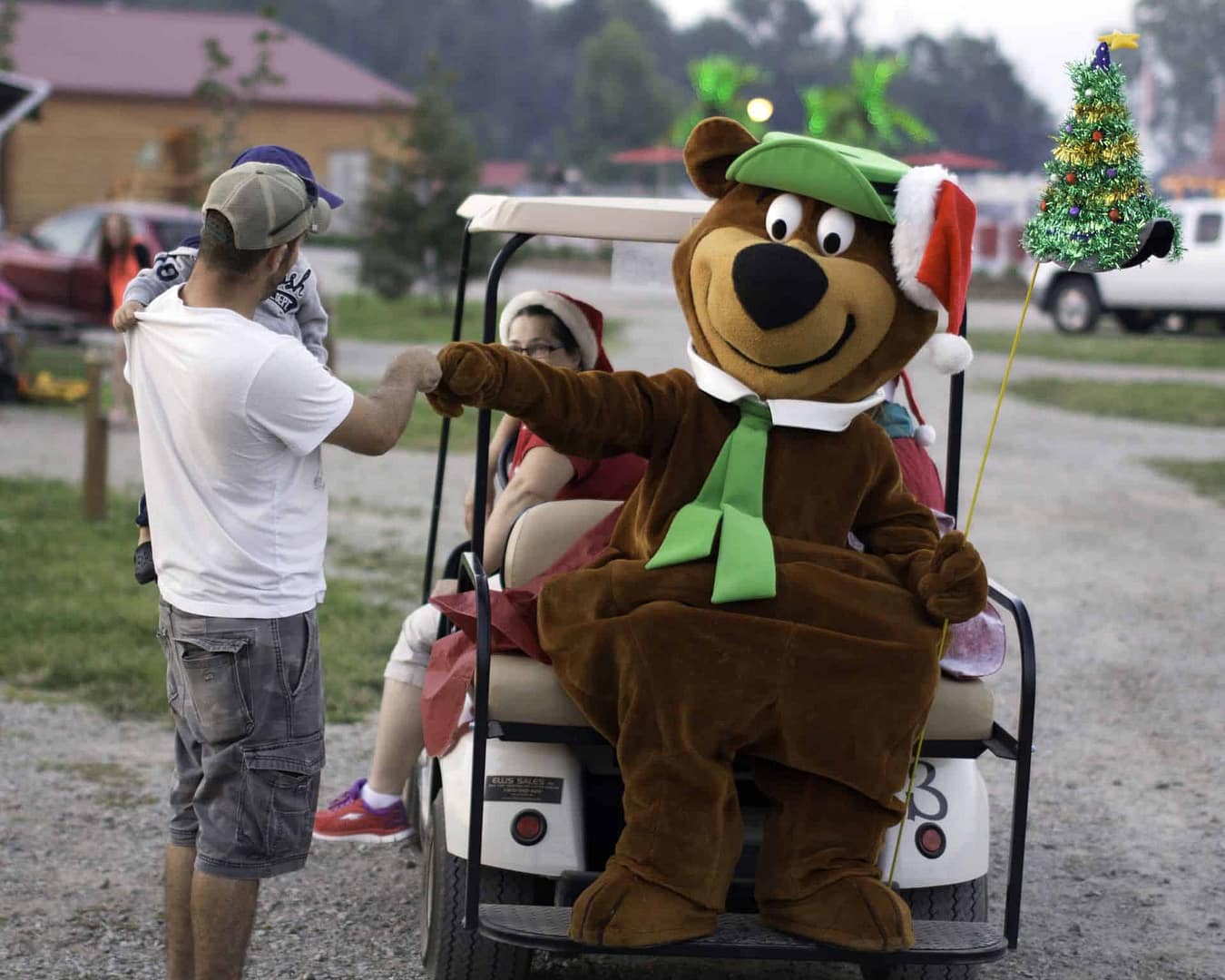 A mascot dressed as a teddy bear is riding in a golf cart at Jellystone Park.