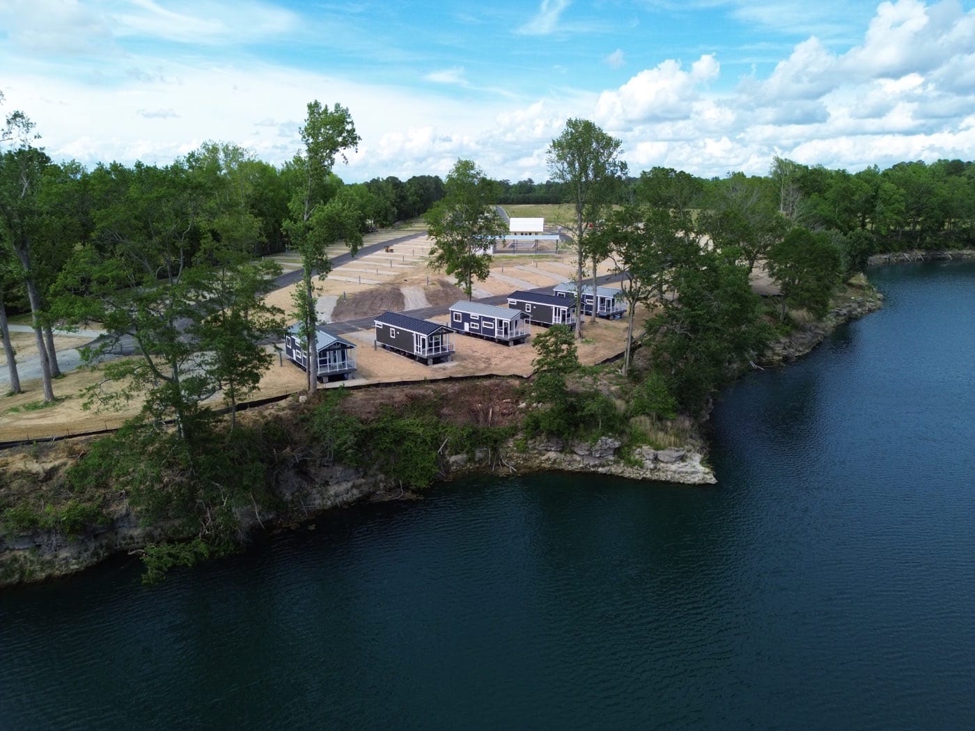 Aerial view of several small houses perched on a cliff overlooking a body of water, with trees surrounding the area and an auto draft construction site in the background.