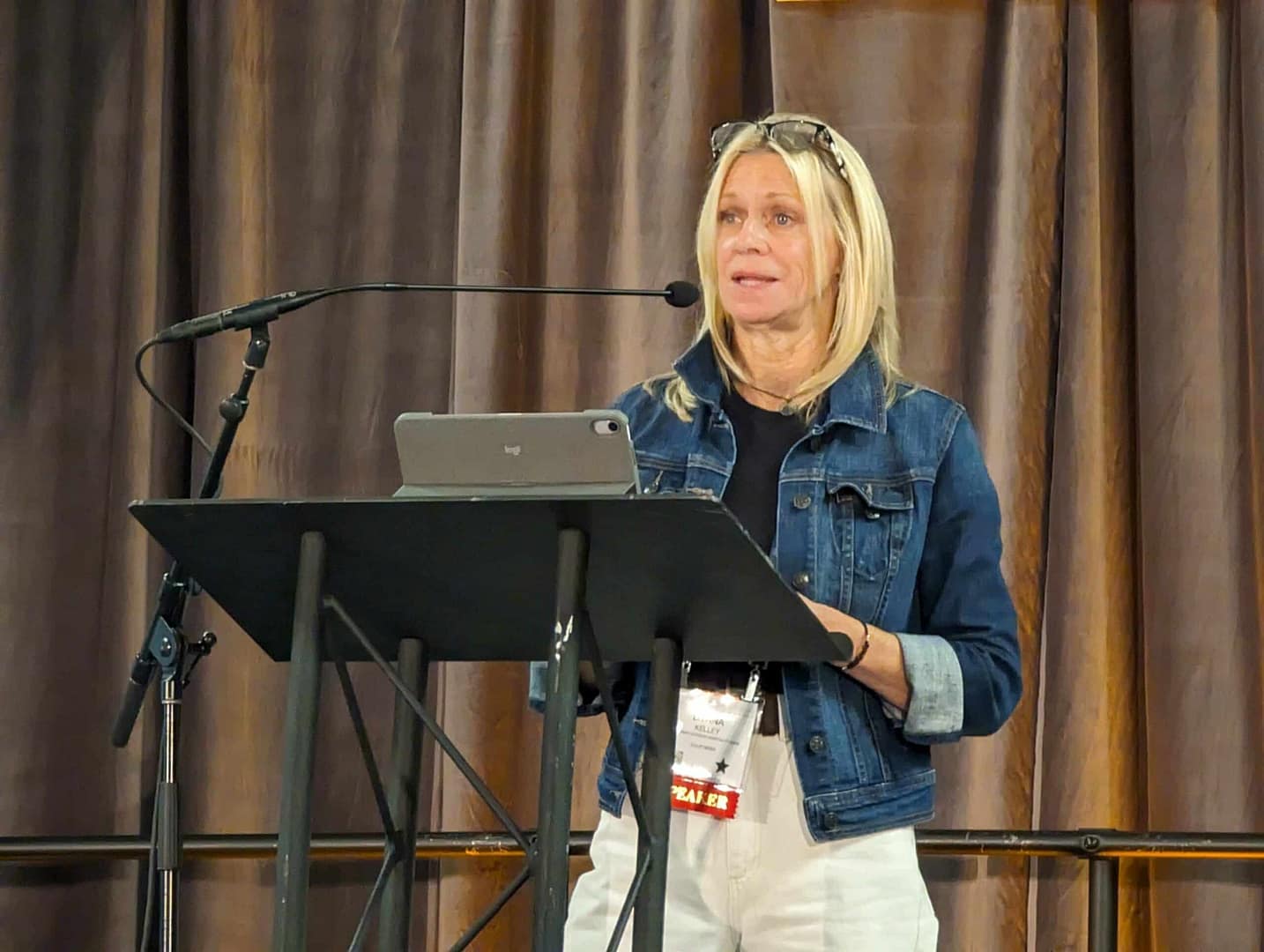 The CalOHA President, wearing a denim jacket, confidently stands at a podium while delivering a speech on Navigating Risk Management at The Glamping Show Americas.