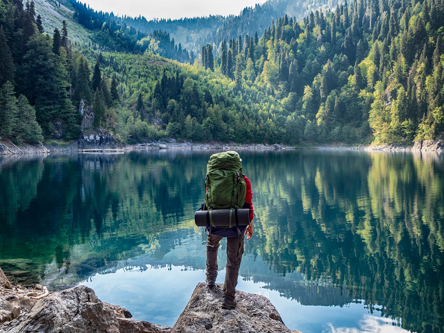 A man with a backpack standing on a rock next to a lake.