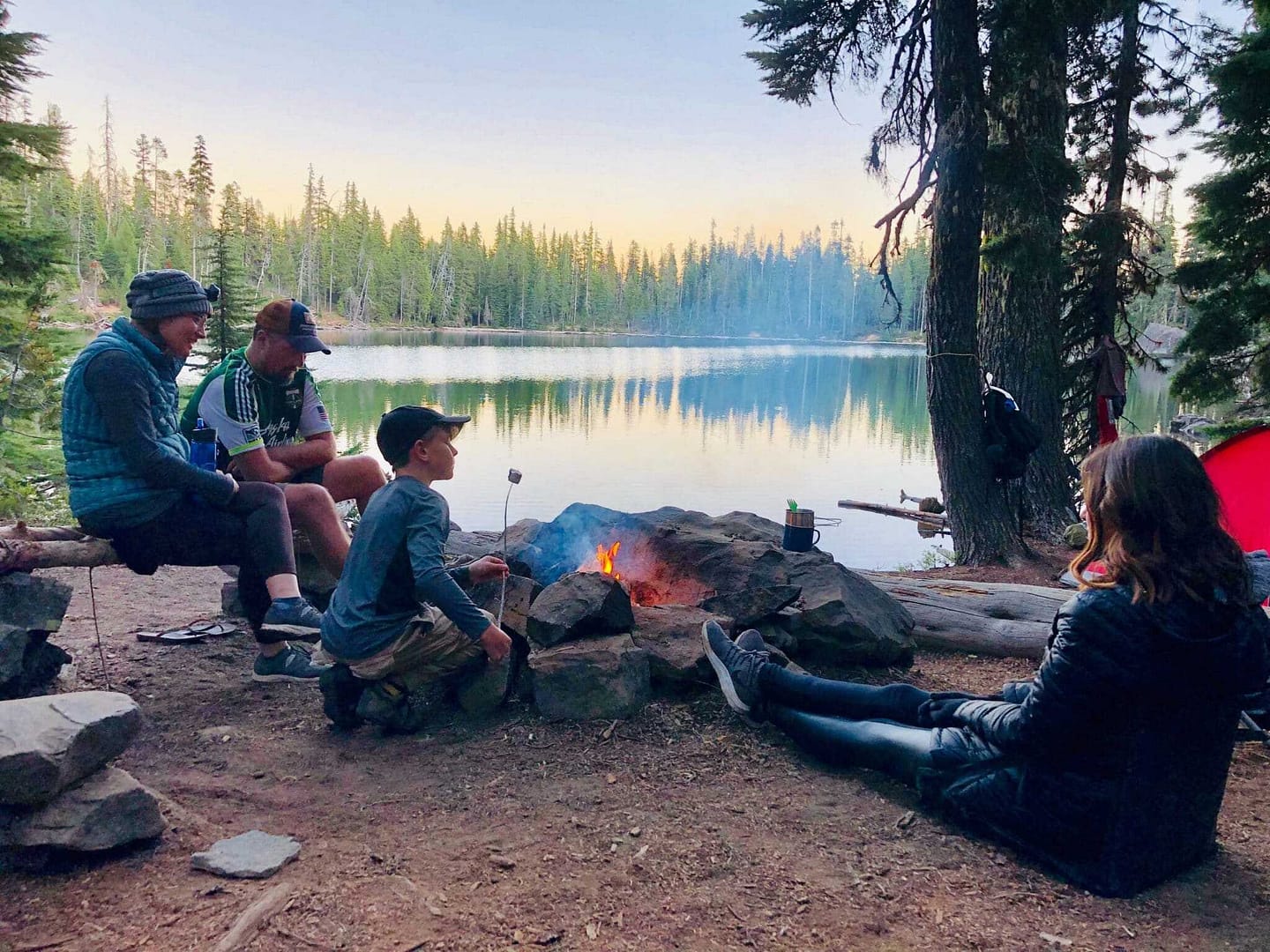 A group of people collaborates around a campfire near a lake.