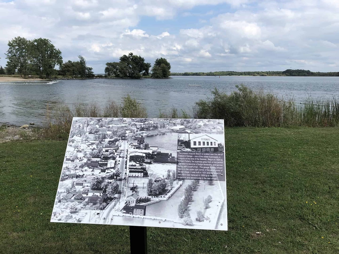 A sign in front of Iroquois Local lake with a boat in the background.