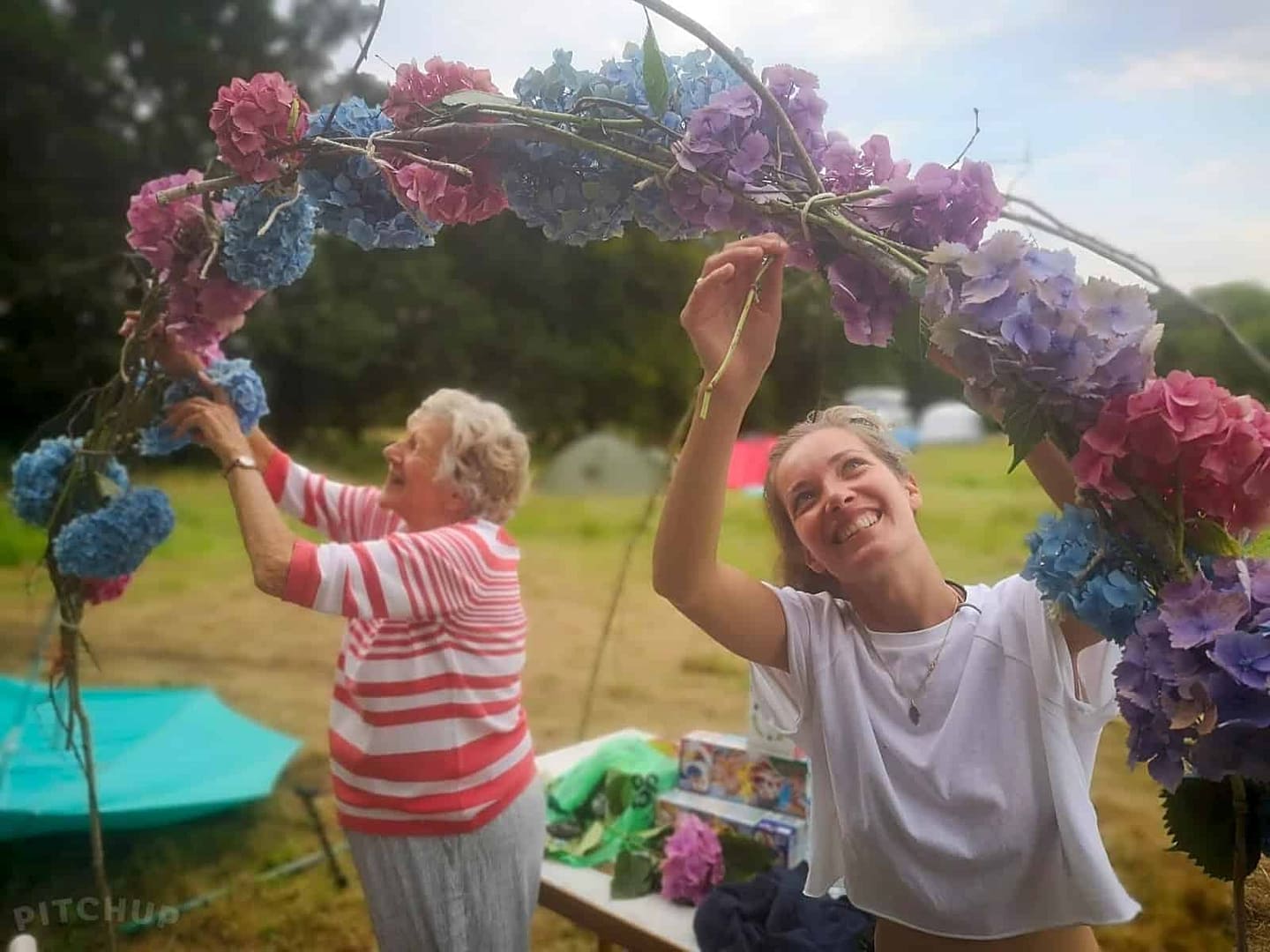 Two women from the UK are making a flower arch at a festival.