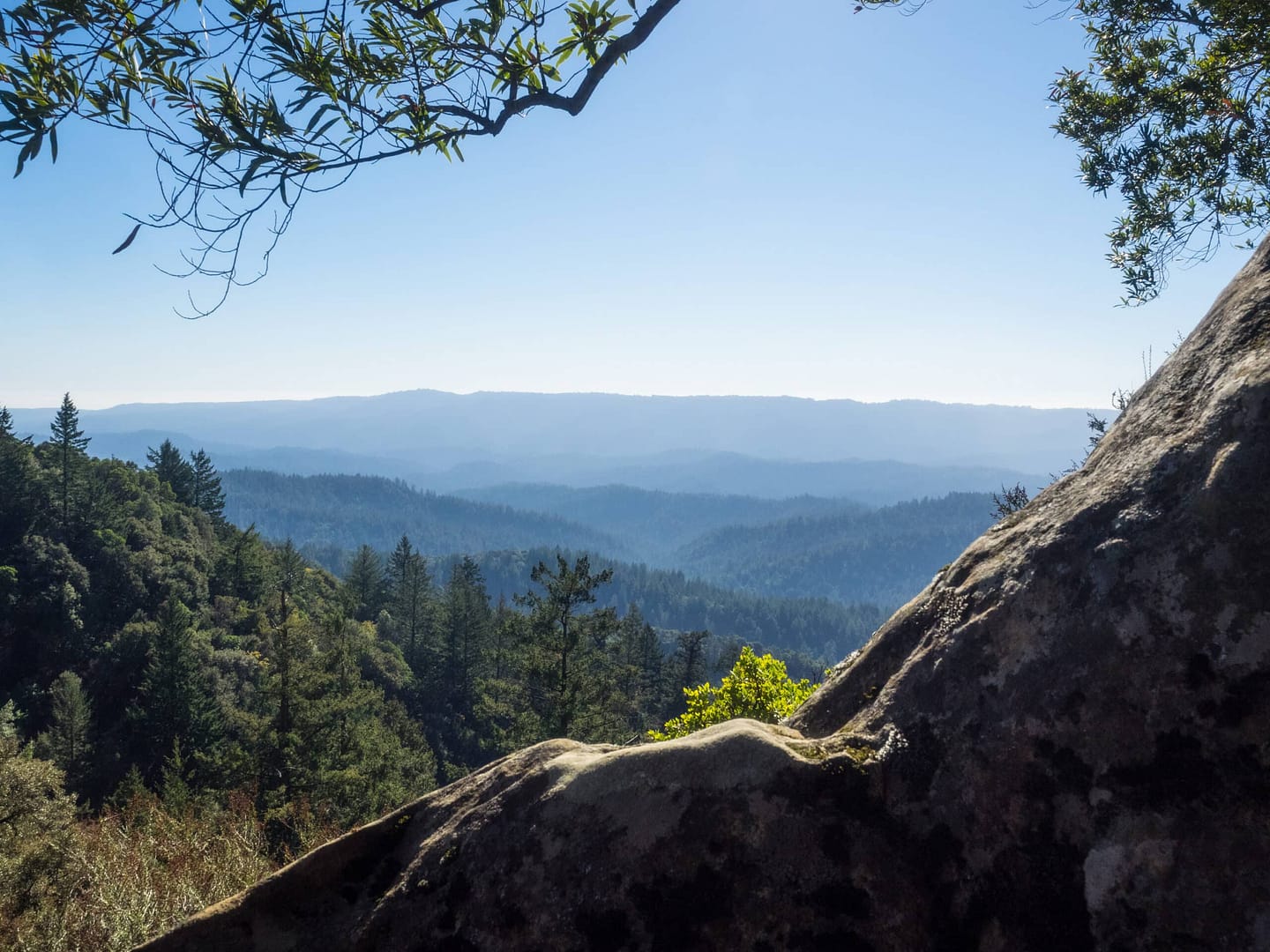 View from a rocky ledge overlooking a vast forest with layers of misty mountains in the distance under a clear sky, capturing the essence of California State Parks.