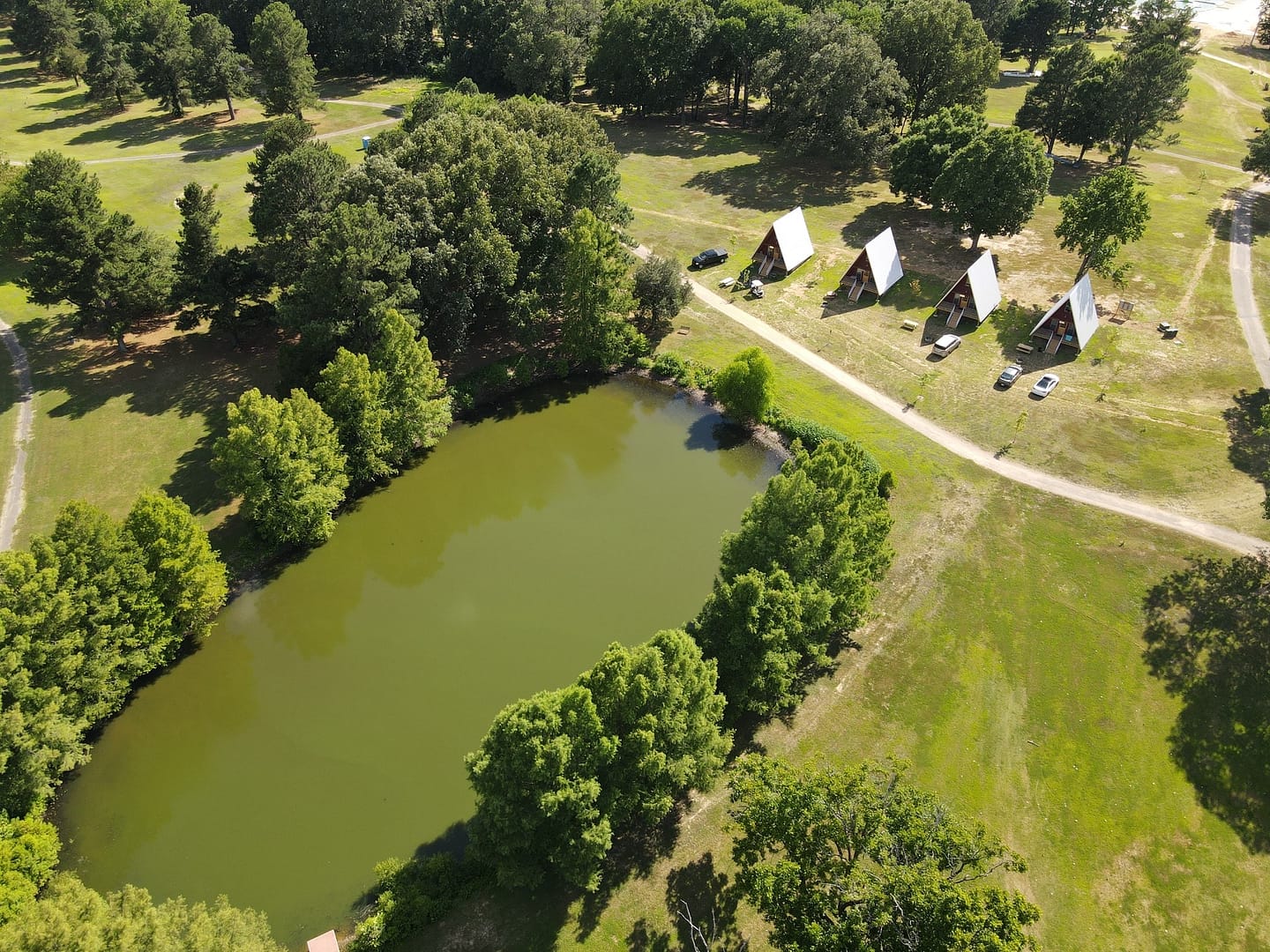 Aerial view of a small lake with triangular cabins and vehicles nearby at Serendipity Resort, surrounded by greenery and pathways.