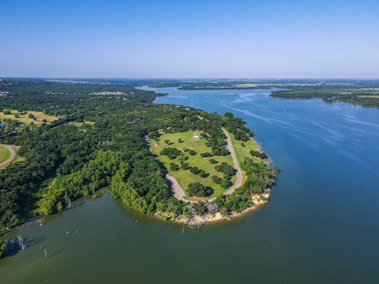 Aerial view of a verdant peninsula with a winding road extending into a large blue Lake Waco, surrounded by lush greenery and scattered clouds overhead.