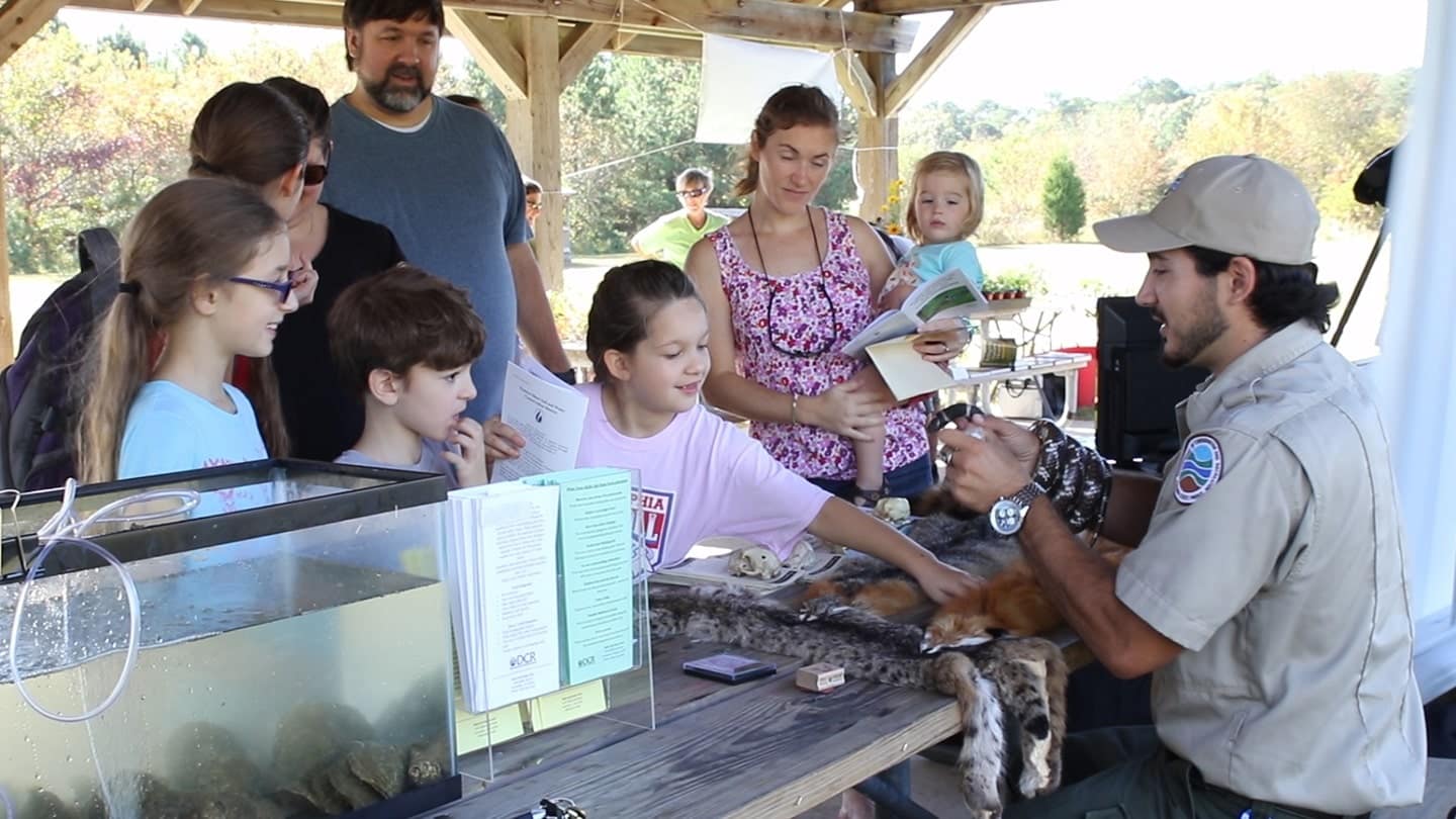 A group of children at Kiptopeke State Park, engaged in an Outdoor Exploration Day with a zoo keeper at a table.
