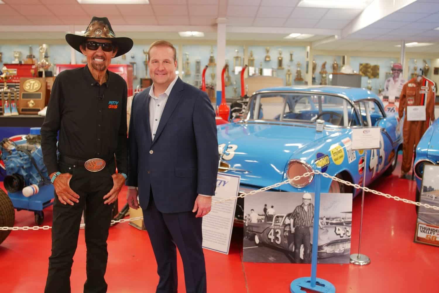 A man in a suit standing next to a car, while another man in a cowboy hat looks on.